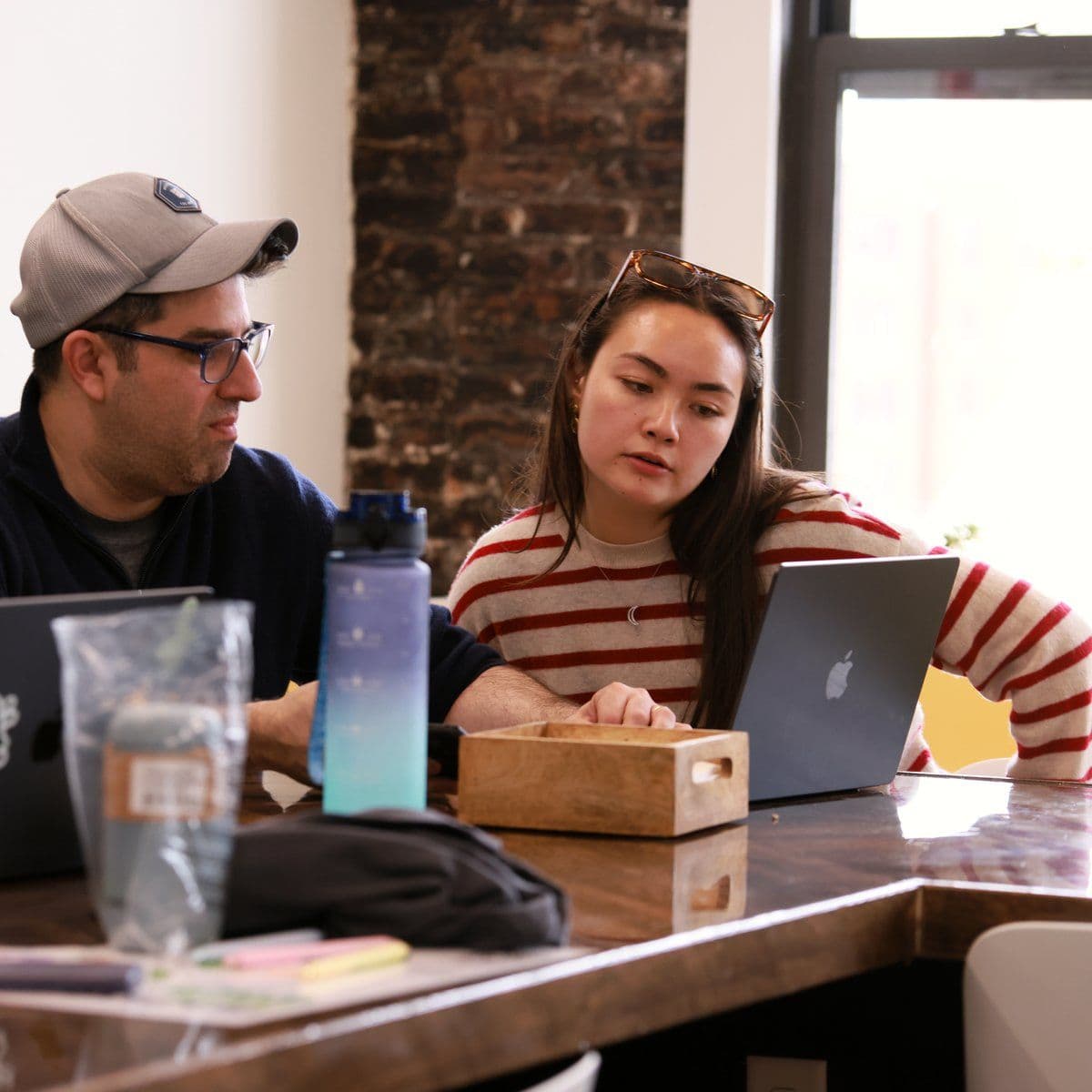 Two teammates work through a problem together on one laptop by the office window